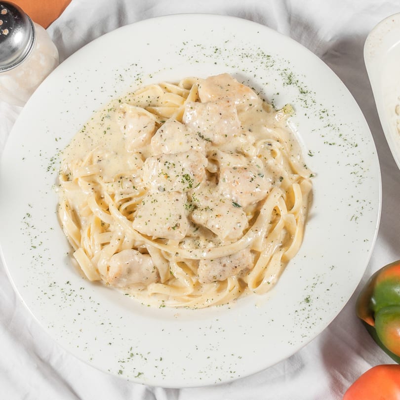 Creamy chicken fettuccine alfredo with parsley flakes on a white plate, with a cheese shaker nearby.